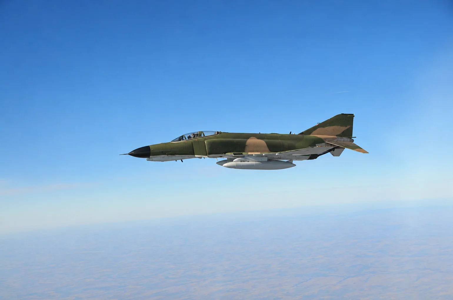Illustrative edit of a camouflaged F-4 Phantom II in flight against a blue sky, with military markings removed