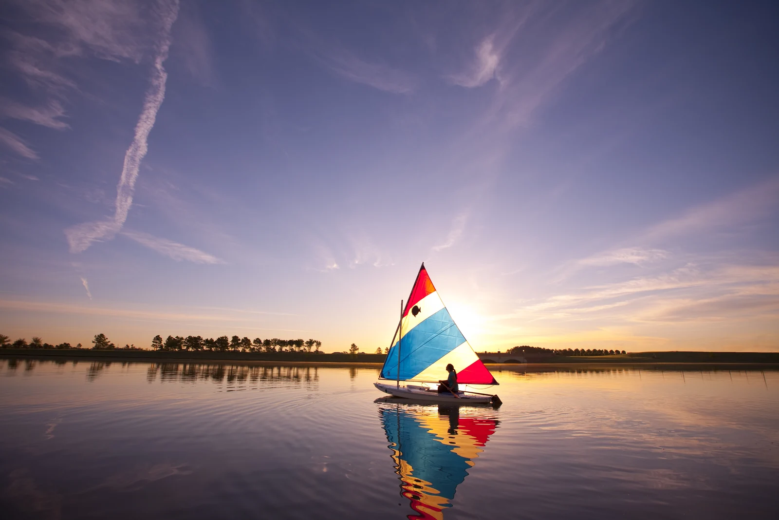 A colorful sailboat reflected in a calm Bridgeland lake under a sunset sky