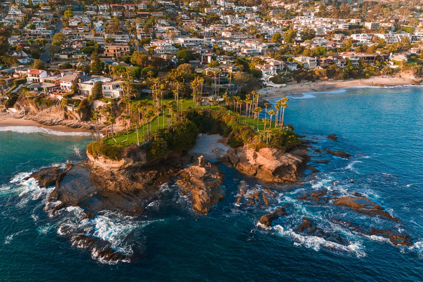 Golden-hour aerial of Laguna Beach coves, palms, coastal homes and rocky promontory