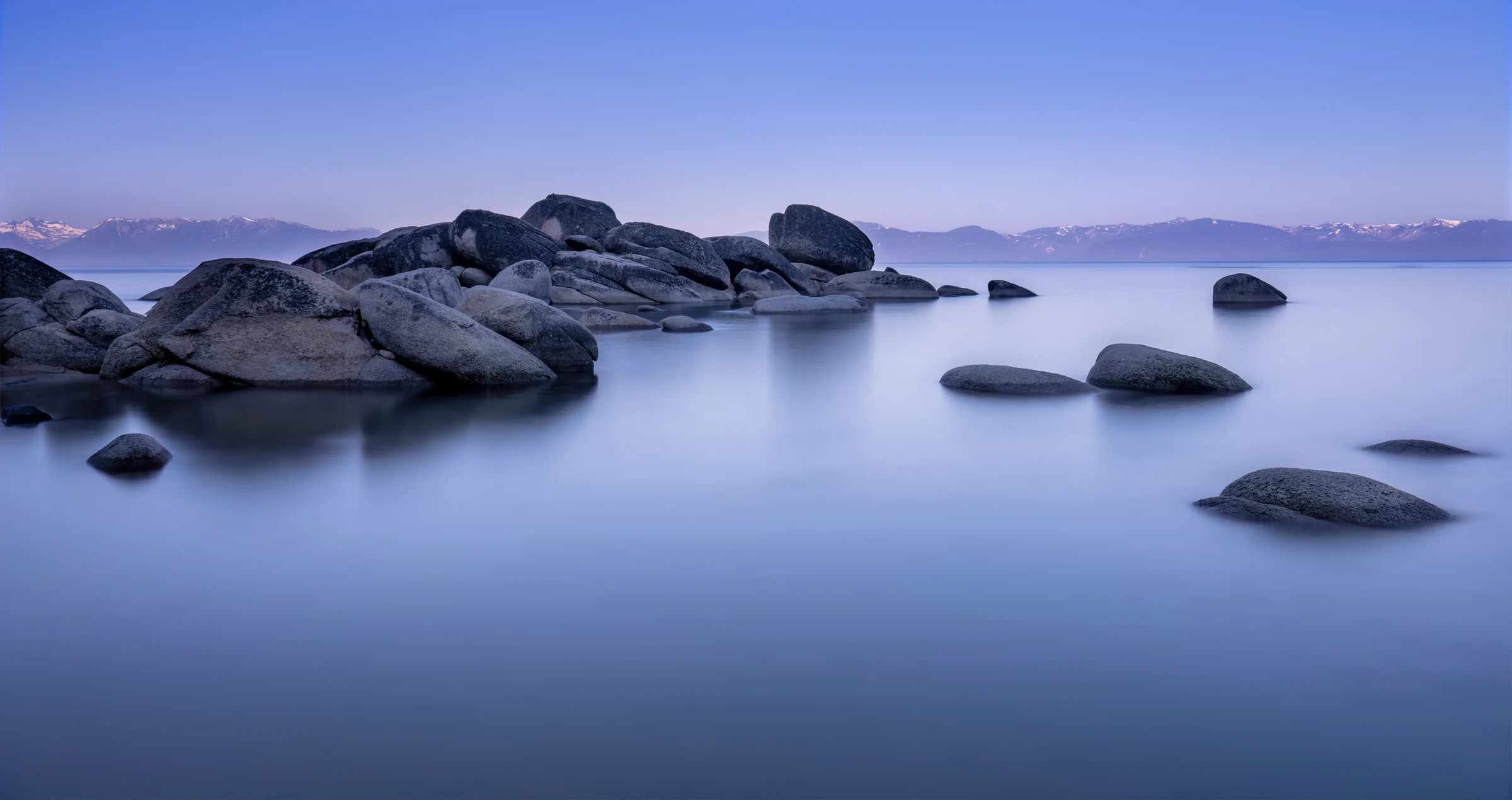 Smooth blue Lake Tahoe water around granite boulders beneath the distant Sierra
