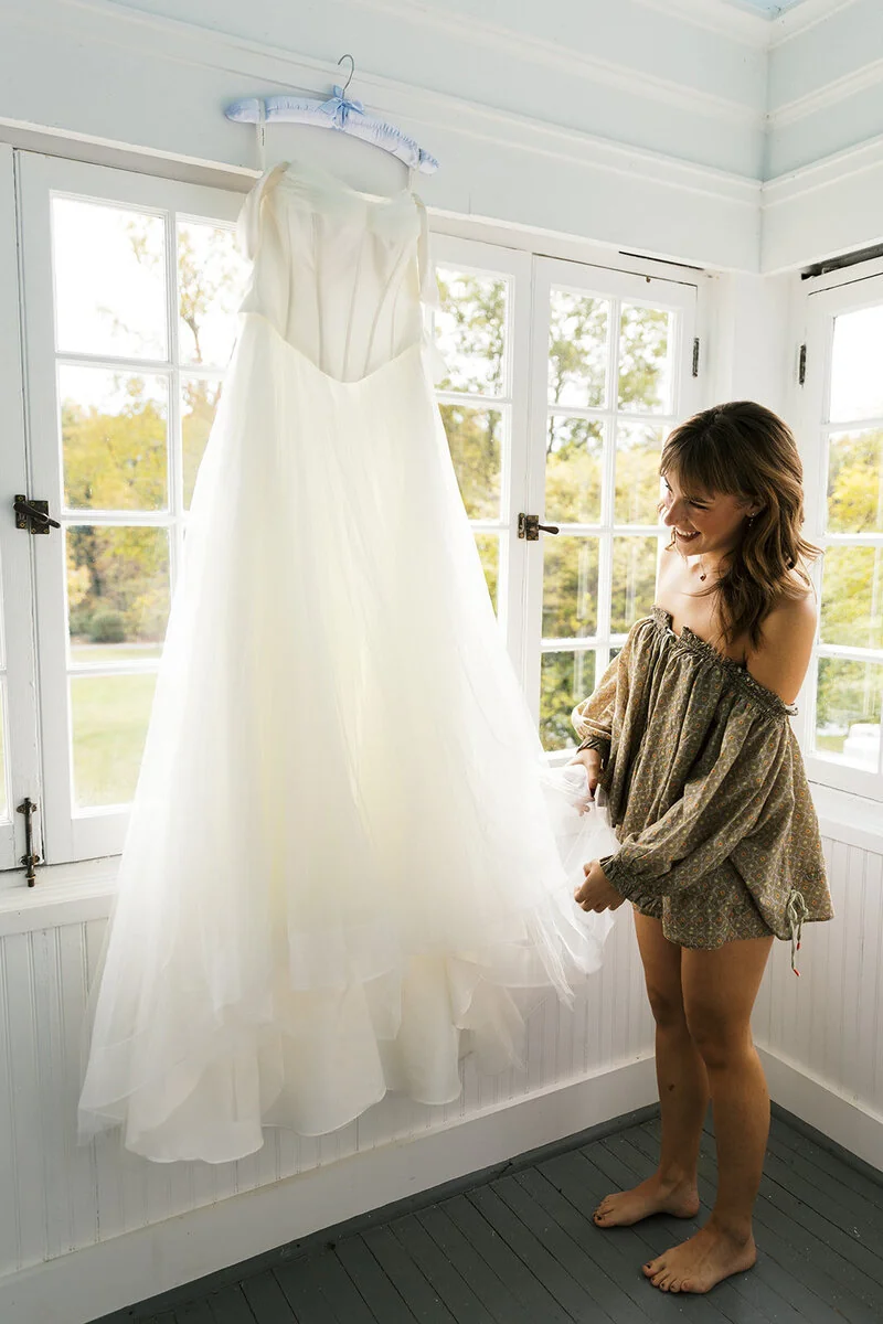 A bride stands beside her hanging wedding dress in a light-filled room.