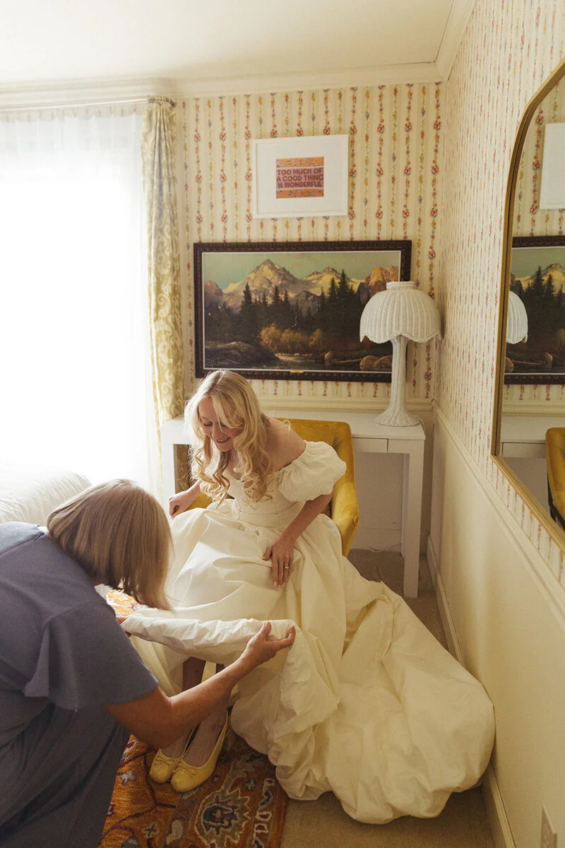 A seated bride smiles as another woman adjusts the skirt of her wedding dress.