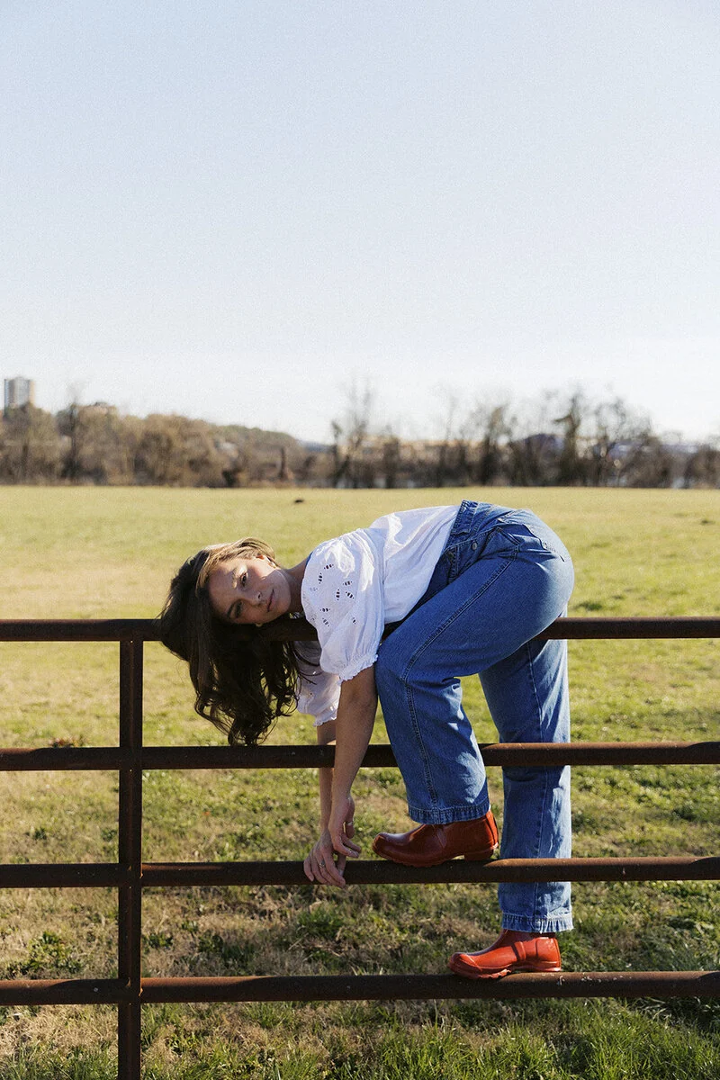 A woman in a white blouse, blue jeans and brown boots poses on a field gate.
