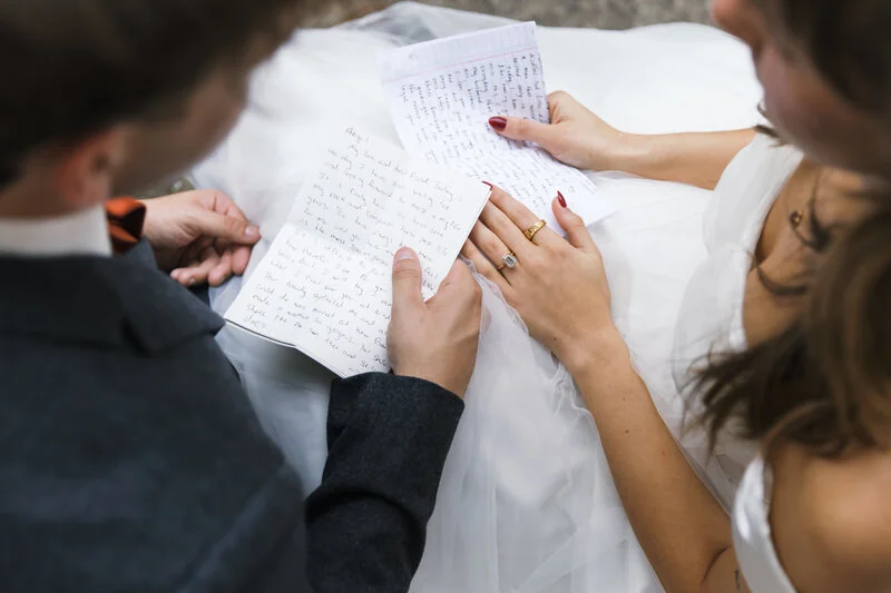 A bride and groom hold handwritten pages, with wedding rings visible.