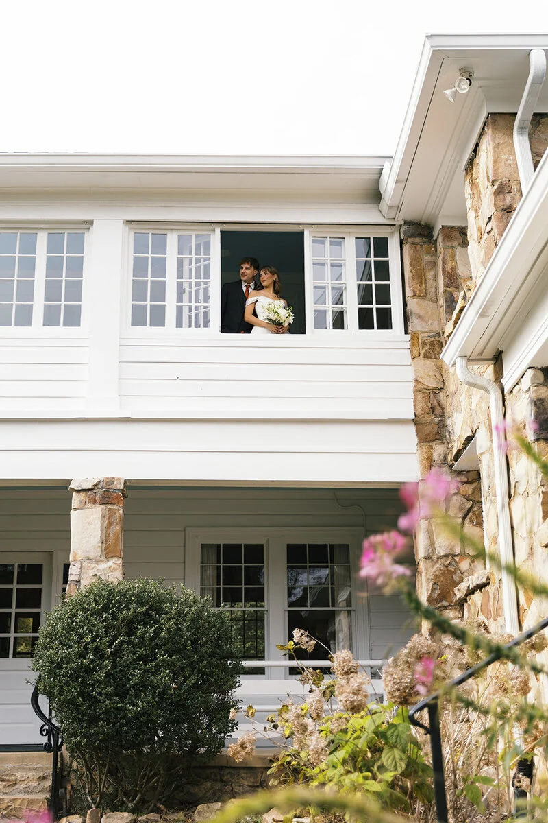 A bride holding flowers and a groom stand together at an open upstairs window.