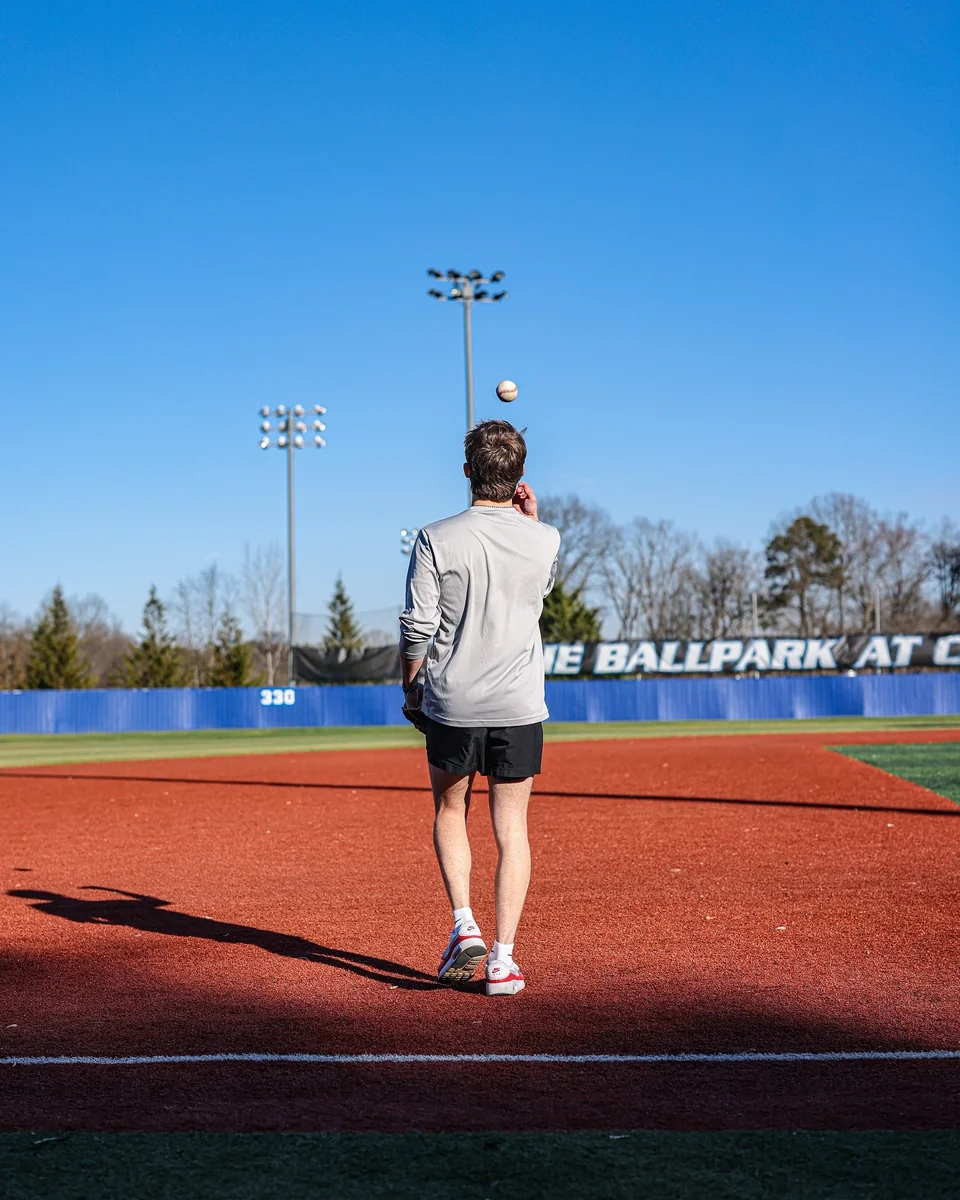 A baseball player seen from behind tosses a ball into the air on a sunlit field.