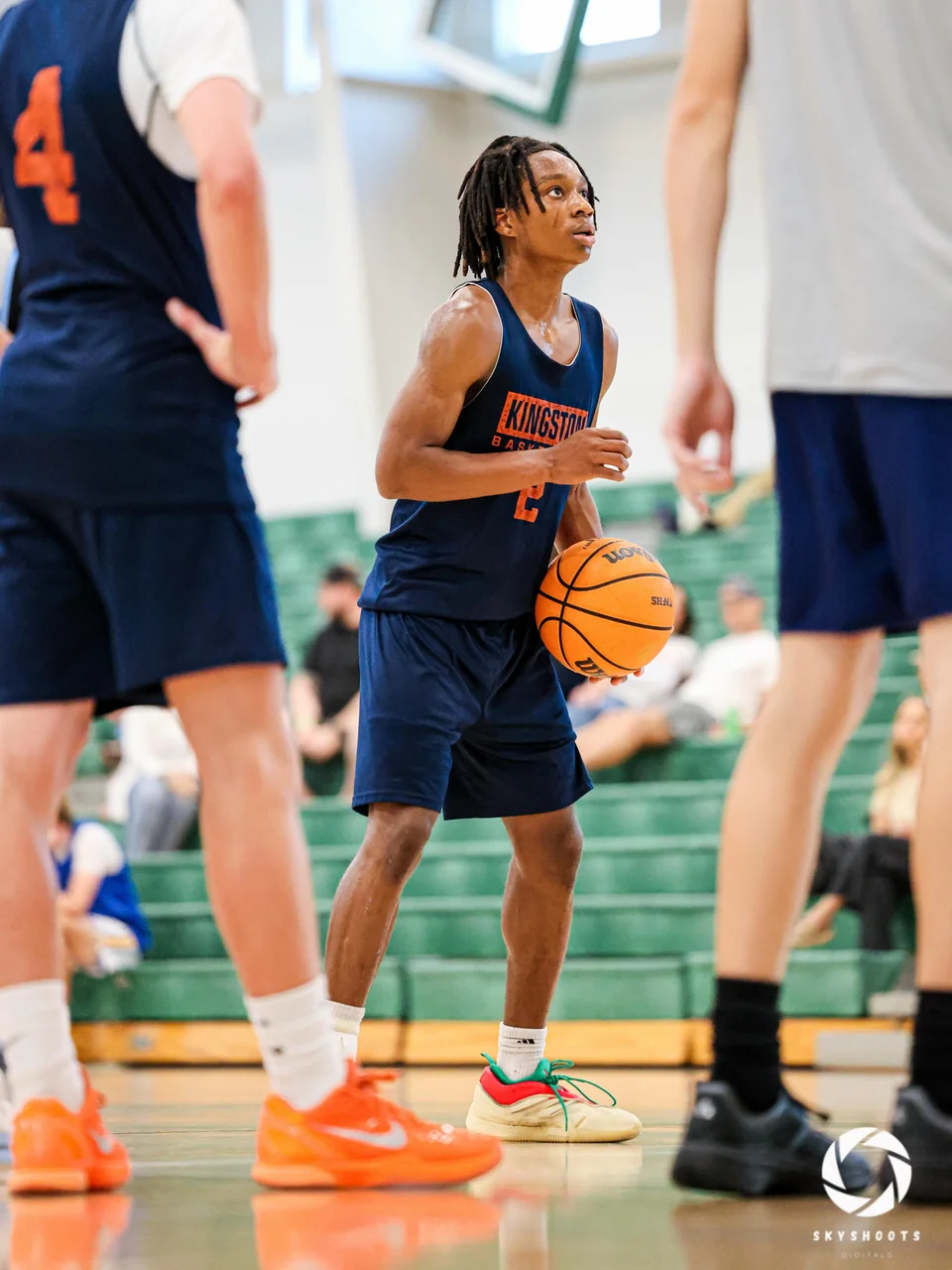 A basketball player in a dark uniform holds the ball while looking toward the basket, framed by other players in a gym.