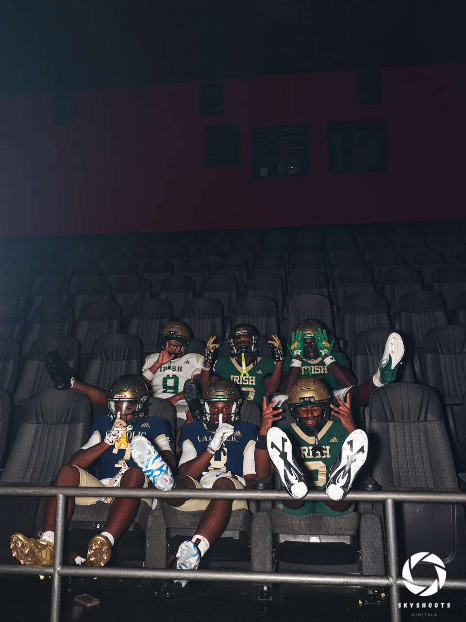 Six football players in helmets and uniforms pose together in rows of theater-style seats in a dark interior.