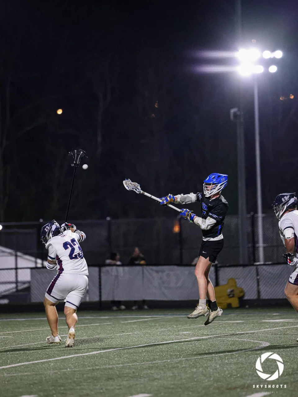 A lacrosse player in a dark uniform jumps while extending a stick during nighttime play.