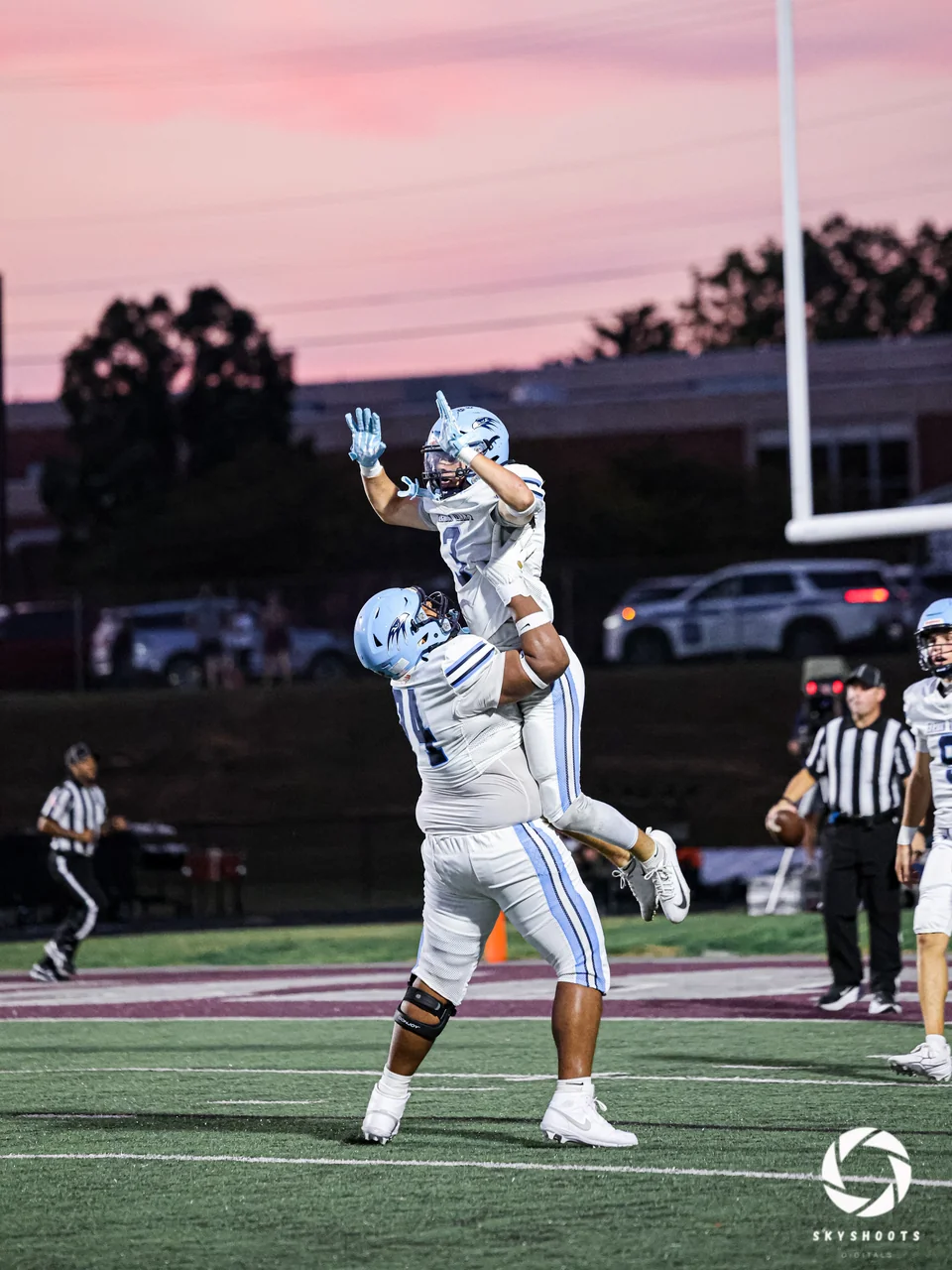 Two football players in light uniforms celebrate beneath a pink evening sky.
