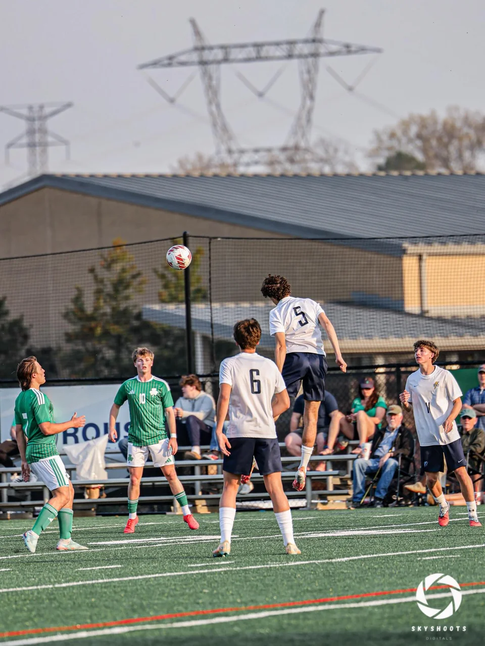 A soccer player in a white shirt leaps toward an airborne ball, with other players nearby.