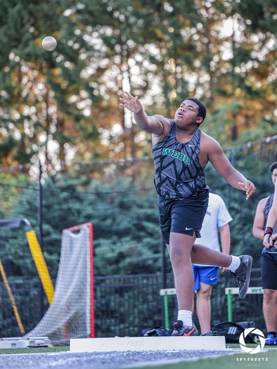 An athlete releases a shot put from the throwing circle.