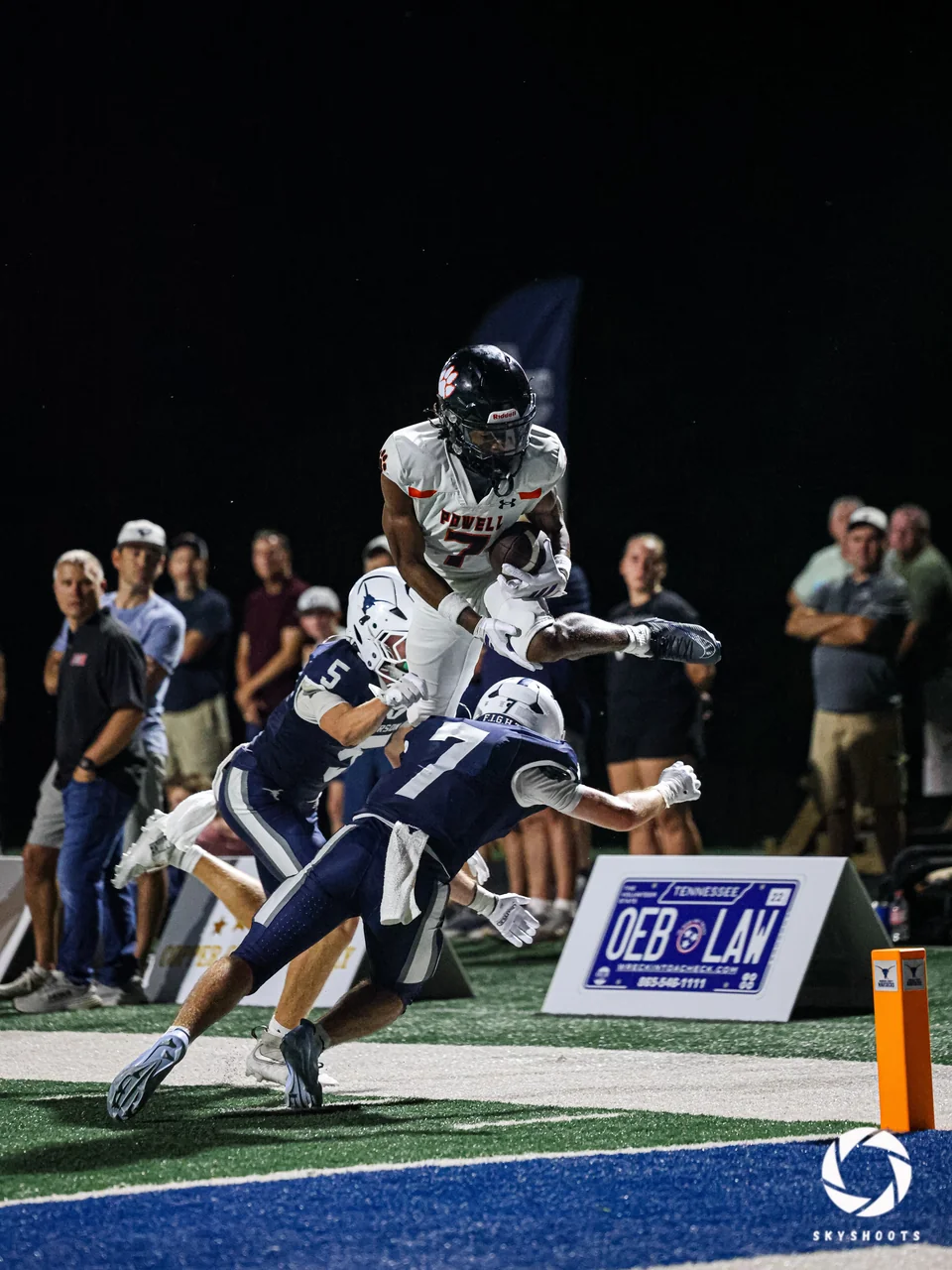 A football runner leaps above defenders near the sideline at night.