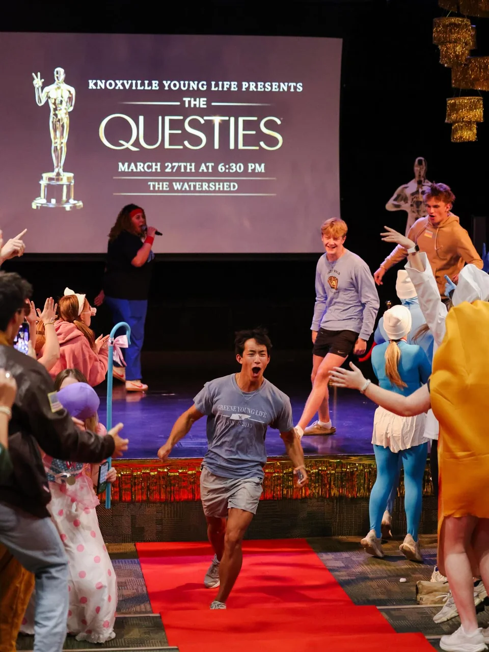 A participant moves along a red carpet between cheering attendees in front of a stage. Projected signage identifies Knoxville Young Life presents The Questies.